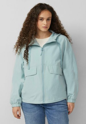 Kobieta z długimi, kręconymi włosami, ubrana w jasnoniebieską kurtkę na zamek z kapturem i sznureczkami oraz niebieskie dżinsy, stojąca na jednolitym tle.