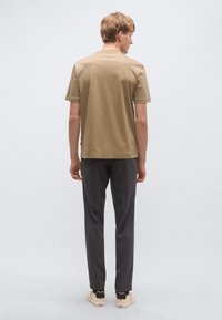 Bézs rövid ujjú póló kerek nyakkivágással, sötétszürke nadrággal és világos tornacipőkkel, hátulnézetből.