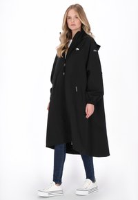 Fekete esőkabát kapucnival, cipzáras záródással, rugalmas mandzsettákkal, oldalzsebekkel és logórészlettel. Kék farmernadrág és fehér tornacipő fölött viselve.