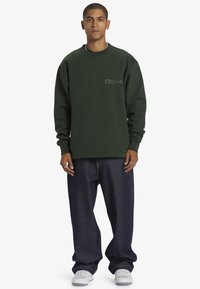 Grön crew-neck sweatshirt med litet grått logotyp, tillsammans med lösa blå jeans och vita sneakers. Modellen står med händerna vid sidorna.