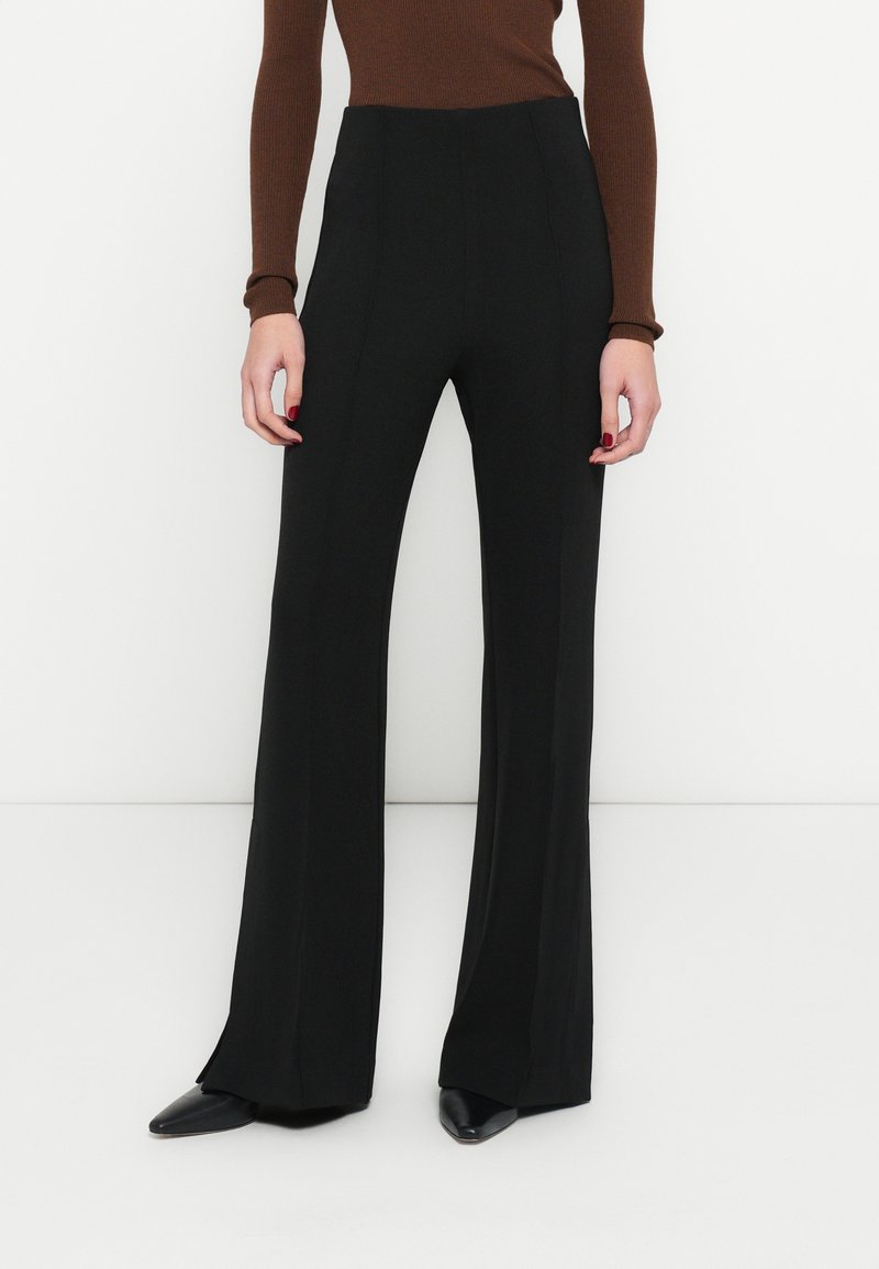 Čierne široké nohavice z hladkého materiálu, s vysokým pásom a prednými záhybmi, v kombinácii s čiernymi lodičkami so špicatou špičkou.