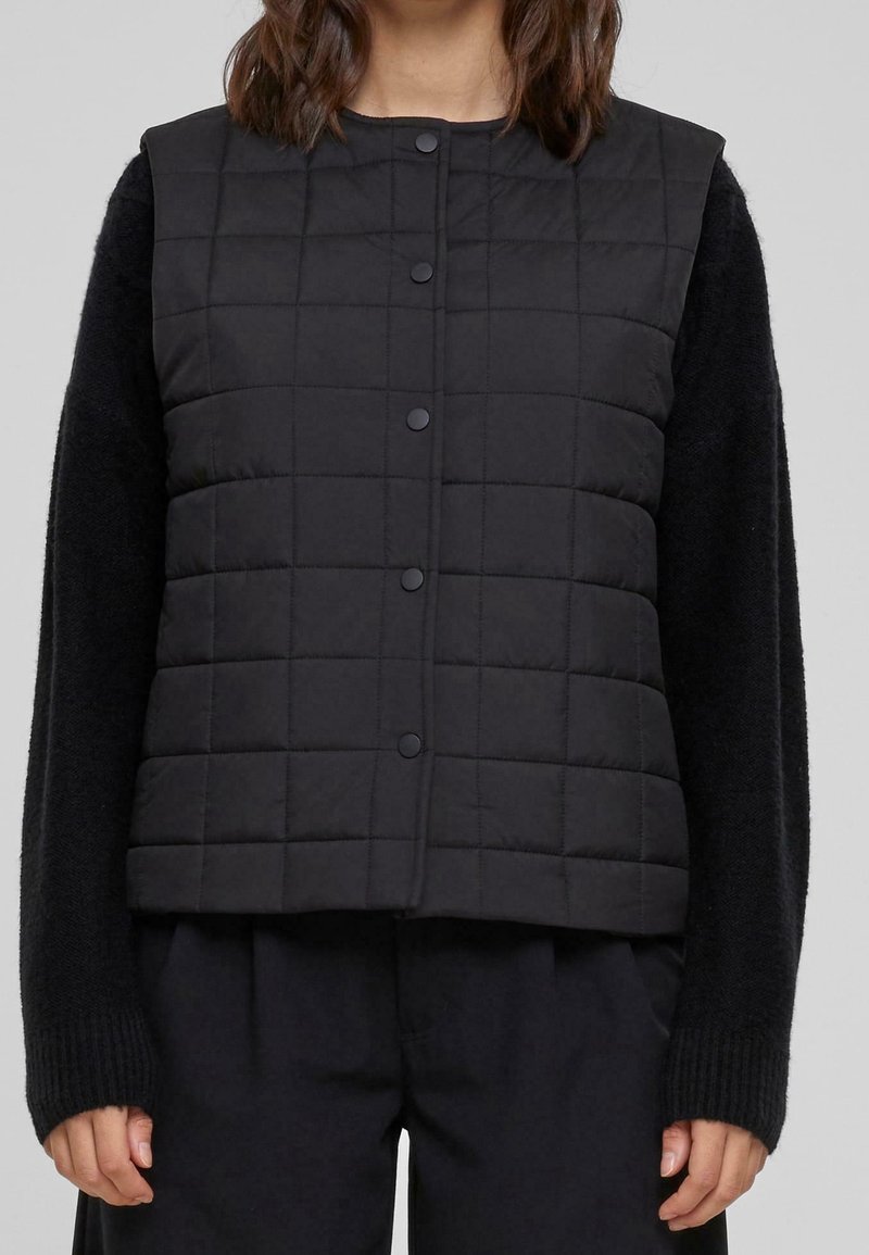 Femme portant une veste matelassée noire avec des boutons-pression par-dessus un pull en maille noir à manches longues et un pantalon noir, les mains détendues le long du corps.