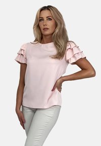 Jasnoróżowa bluzka z falbanami, krótkimi rękawami, miękkiego materiału, okrągłym dekoltem i luźnym krojem, zestawiona z dopasowanymi jasnymi spodniami.