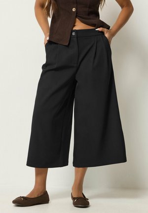 Femme portant un pantalon noir large et court, un top marron sans manches et des ballerines marron, debout les mains dans les poches.