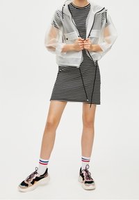 Klare, leichte, taillierte Jacke aus transparentem Stoff über einem schwarz-weiß gestreiften Kleid. Kombiniert mit klobigen Turnschuhen und gestreiften Socken.