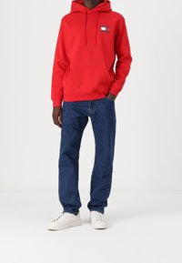 Felpa rossa con tasca frontale e cappuccio, con un patch logo. Abbinata a jeans blu e sneaker bianche.