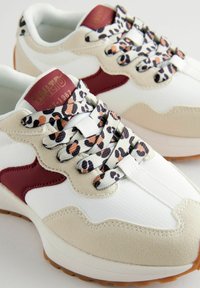 Sneakers z białą siateczką na górze, beżowymi zamszowymi akcentami, bordowym logo i sznurówkami w wzór lamparta. Gruba gumowa podeszwa z jasnobrązowym bieżnikiem.