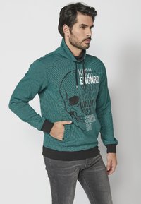 Teal dukserica s kapuljačom, teksturiranog uzorka, crnim manžetama i prednjim džepom. Sadrži grafiku lubanje i tekst u kontrastnim bojama.