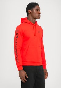 Hombre vistiendo una sudadera brillante roja de Emporio Armani con bolsillo frontal y texto del logo en el pecho y la manga, combinada con pantalones negros.