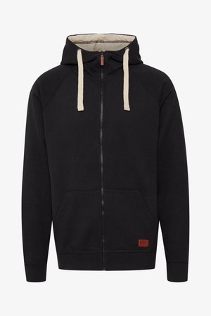 Sudadera con cremallera negra con cordón crema, bolsillos frontales, mangas raglán, puños acanalados y una etiqueta marrón texturizada en la parte inferior derecha.