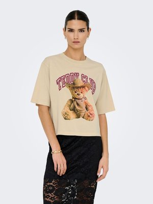 Vrouw draagt een beige oversized T-shirt met teddybeer en de tekst "Teddy Club", gecombineerd met een zwarte kanten rok en een gouden armband.