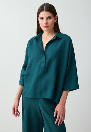 Giovane donna con lunghi capelli castani che indossa orecchini a cerchio dorati e una camicia azzurro petrolio morbida con bottoni con pantaloni abbinati su uno sfondo semplice.