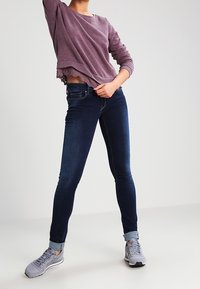Jeans skinny azul oscuro con una cintura de altura media, combinados con un sudadera corta de color púrpura claro que presenta un dobladillo de encaje. Zapatillas deportivas grises.
