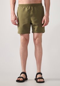 Green swim shorts with an elastic waistband, smooth texture, and side pockets. Worn with black sandals featuring adjustable straps.