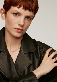 Abrigo de cuero negro con solapas anchas; collar de cadena dorada; mano adornada con un anillo de piedra azul; presenta piel clara y cabello corto rojo.