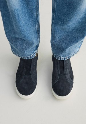 Des chaussures noires en daim à enfiler, portées avec un jean bleu clair délavé, se tenant sur un sol de couleur claire.