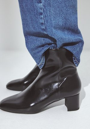 Personne portant des bottines noires brillantes à petits talons carrés, avec un jean en denim bleu couvrant le dessus des bottines.