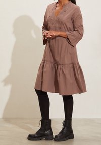 Femme portant une robe mauve jusqu'aux genoux avec des détails brodés, des collants noirs et des bottes de combat à lacets noires, se tenant contre un mur uni.