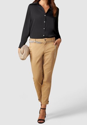 Frau trägt schwarze Bluse mit Knöpfen, beigen Hosen mit silbernem Kettengürtel, hält eine gewebte Clutch und trägt schwarze Sandalen mit Absatz und Fußkettchen.