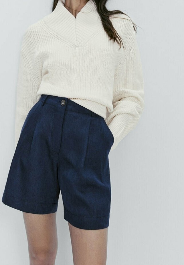 Pull en maille crème avec un col montant et un motif diagonal, associé à un short taillé bleu marine avec des plis et des poches latérales.