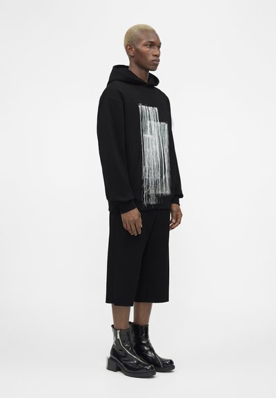 Moški s kratkimi svetlo blond lasmi, ki nosi črn pulover s kapuco z belimi cofki, črne široke kratke hlače in sijoče črne gležnjarje, stoji pokončno od strani.