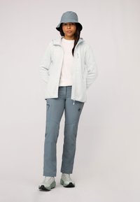 Giacca in pile azzurro chiaro, camicia bianca, pantaloni outdoor grigi con tasche e cappello a secchiello grigio. Presenta un tessuto leggero e un design sportivo.