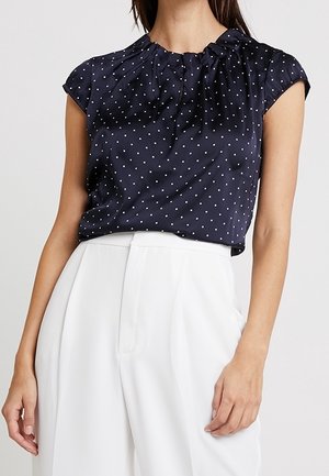 Marineblauwe mouwloze blouse met witte stippen, gecombineerd met wit hoge taille wijde broek op een staand persoon.