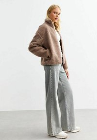 Bruine fleecejas met een hoge kraag, gedragen over een witte top en gecombineerd met lichtblauwe wijdgesneden jeans en witte schoenen.