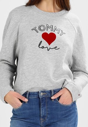 Kobieta ubrana w szarą bluzę z tekstem "TOMMY", czerwoną grafiką serca i napisem "love" w stylu kursywy, w połączeniu z niebieskimi dżinsami, ręce w kieszeniach.