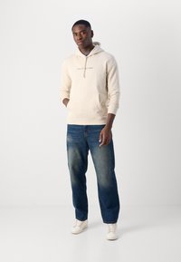 Felpa con cappuccio color beige chiaro con tasca a canguro frontale, abbinata a jeans in denim blu con un leggero sbiadimento. Sneakers bianche completano l'outfit.