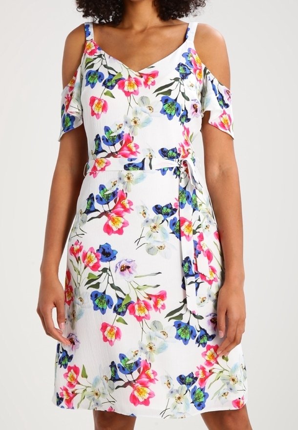 Femme portant une robe blanche à épaules dénudées avec un motif floral rose, bleu et vert et une ceinture nouée à la taille.