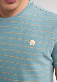 Chemise bleu clair à manches courtes avec des rayures horizontales beiges. Un patch de logo rond est placé sur le côté gauche de la poitrine. Tissu texturé.
