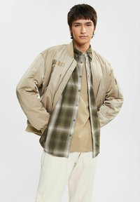 Beige bomber jacket with ribbed cuffs, layered over a green plaid shirt. The shirt is worn over a tan base layer, paired with cream-colored pants.