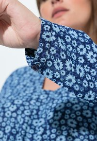 Damska koszula z długim rękawem w kwiatowy wzór stokrotek w kolorze niebieskim i białym. Posiada mankiety na guziki oraz okrągły dekolt. Miękka w dotyku tkanina.