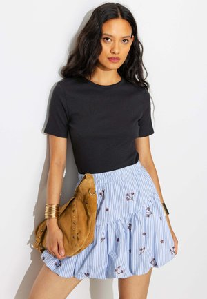 Jeune femme portant un haut noir à manches courtes, une jupe bleue rayée avec un imprimé floral, tenant un sac à main en daim marron et portant des bracelets en or.