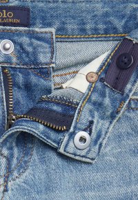 Jeans in denim con un lavaggio azzurro chiaro, cerniera argentata e bottone di rame. Presentano cuciture gialle e un'etichetta di tessuto blu navy.