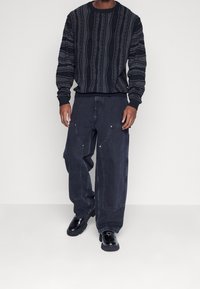 Černá vzorovaná pletená mikina s dlouhými rukávy, doplněná volnými černými džínovými kalhotami s velkými kapsami a stříbrnými kovovými doplňky.