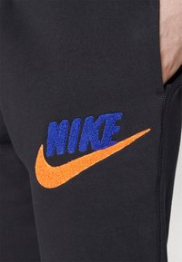 Svarta sweatpants med en blå broderad "NIKE"-logotyp och en orange swoosh. Tyget verkar mjukt och texturerat.