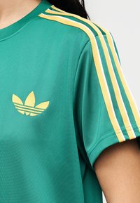 Πράσινο κοντομάνικο μπλουζάκι από απαλό ύφασμα, με κίτρινες και λευκές ρίγες στους ώμους και κίτρινο κεντημένο λογότυπο στο μπροστινό μέρος.