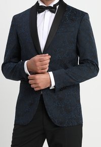 Uomo che aggiusta il polsino di un giacca smoking a righe blu scuro con reversal neri, camicia bianca, cravatta a farfalla nera e pantaloni neri.