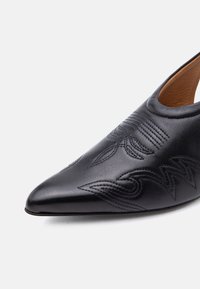 Chaussure en cuir noire à bout pointu avec des motifs cousus complexes et un design bride arrière, présentée sur un fond clair uni.