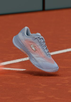 Lichtblauwe tennisschoen met oranje accenten en een wit krokodillenlogo die rode kleideeltjes op een tennisbaan opstrooit.