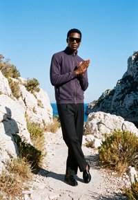 Homme portant des lunettes de soleil, une veste sombre et un pantalon noir, debout les mains jointes sur un sentier rocheux entre des falaises près de la mer sous un ciel bleu clair.