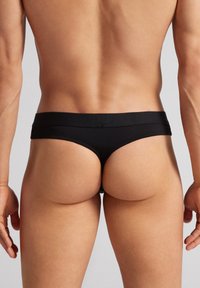 String noir pour hommes en tissu lisse, avec une large ceinture et une couverture minimale, mettant en valeur un design épuré et une forme épousant les contours.