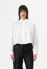 White cropped shirt with a button-down collar and long sleeves, featuring subtle stitching details. Paired with textured black pants.