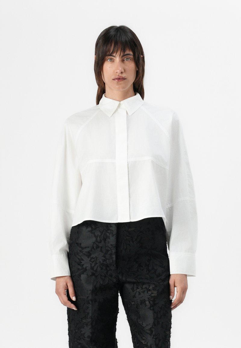 White cropped shirt with a button-down collar and long sleeves, featuring subtle stitching details. Paired with textured black pants.