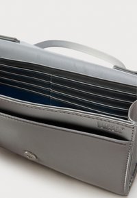 Portefeuille synthétique gris à texture lisse, doté de plusieurs emplacements pour cartes, d'un intérieur bleu et d'une fermeture à bouton en argent. Nom de la marque visible.