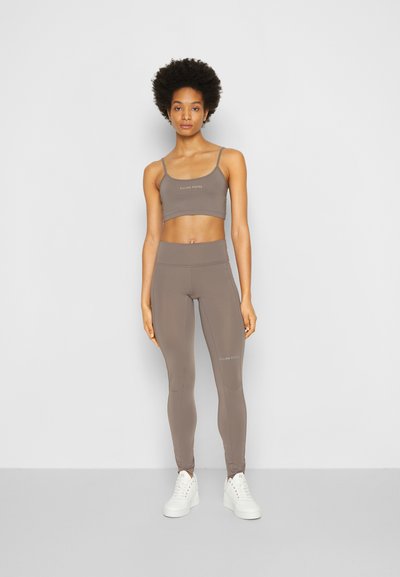 Sutiã desportivo castanho e leggings de cintura alta feitos de um tecido elástico, apresentando uma marcação mínima de texto. Ténis brancos completam o conjunto.