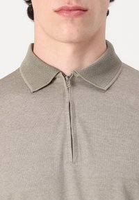 Svetlo siva polo majica s zatvaranjem na zip na vratu in teksturiranim mrežastim ovratnikom. Tkanina ima gladek, strukturiran videz.