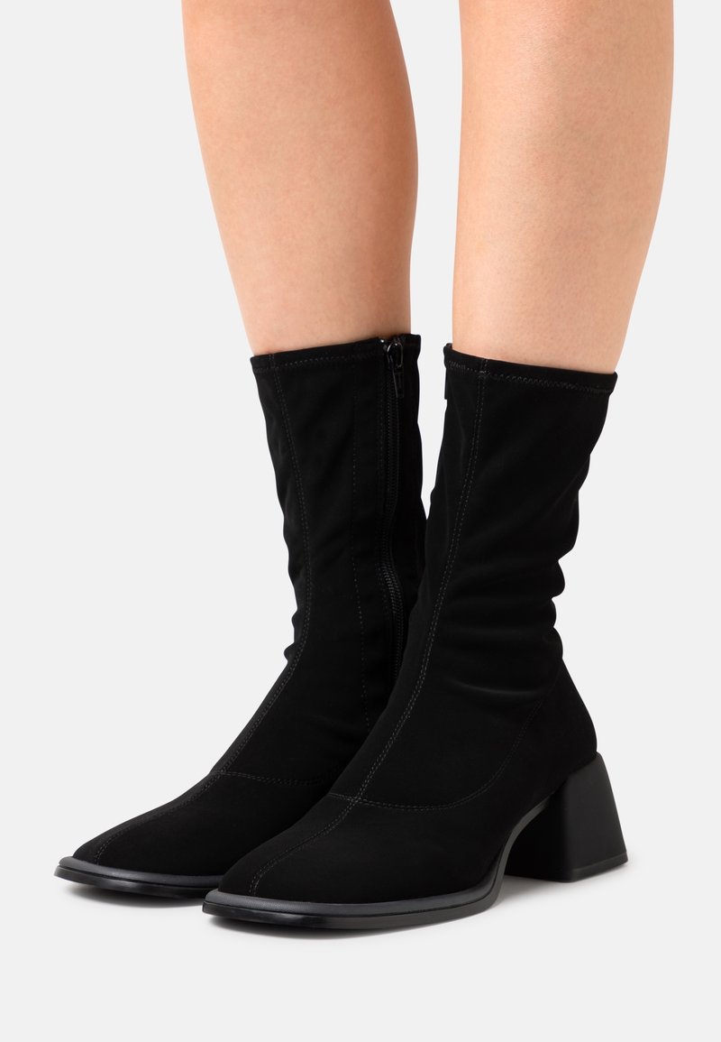 Bottes noires mi-mollet avec talons bloc et fermetures éclair latérales portées sur jambes nues sur un fond clair uni.